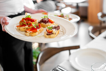 Waiter serving bruschetta topped with tomatoes, greens, and meat in cozy restaurant setting,...