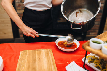 Close-up photo of cook, waiter young woman pouring Ukrainian fresh red borscht from big pot with...