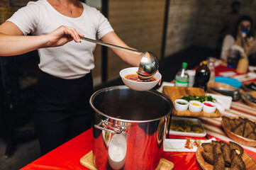 Photo of cook, waiter young woman pouring Ukrainian borscht from a large pot into a plate in a...