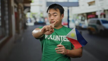Young man outdoors holding philippines flag in urban street setting wearing green volunteer shirt expressing gesture of awareness and engagement