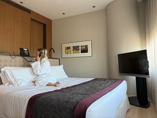 Woman in white hotel bathrobe talking on a landline phone while sitting on the bed. Possibly making...
