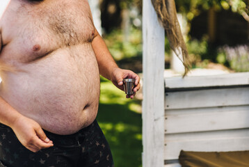 Fat, obese drunk Russian man in summer on vacation, party holds metal shot glass with vodka in hand...