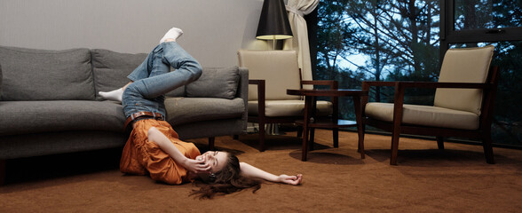 Young woman in casual orange top and jeans, showing relaxed emotions while lying on the sofa in a...
