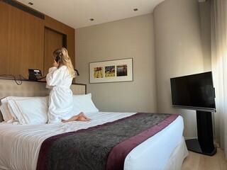 Woman in white hotel bathrobe talking on a landline phone while sitting on the bed. Possibly making...