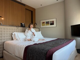 Woman in white hotel bathrobe talking on a landline phone while sitting on the bed. Possibly making...