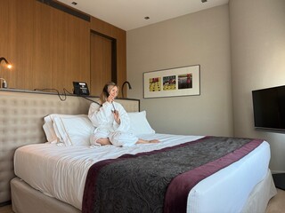 Woman in white hotel bathrobe talking on a landline phone while sitting on the bed. Possibly making...