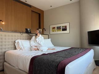 Woman in white hotel bathrobe talking on a landline phone while sitting on the bed. Possibly making...
