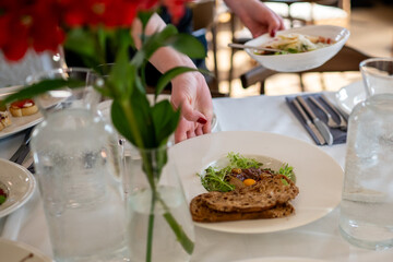 Elegant restaurant table setting with gourmet toast and greens on a white plate, served by hand, surrounded by glassware, cutlery, and red flower centerpiece.