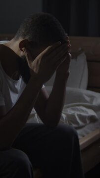 Black bearded man cries hiding face in palms on messy bed