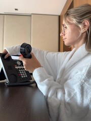 Woman in white bathrobe standing in a hotel room and talking on a landline phone. Possibly...