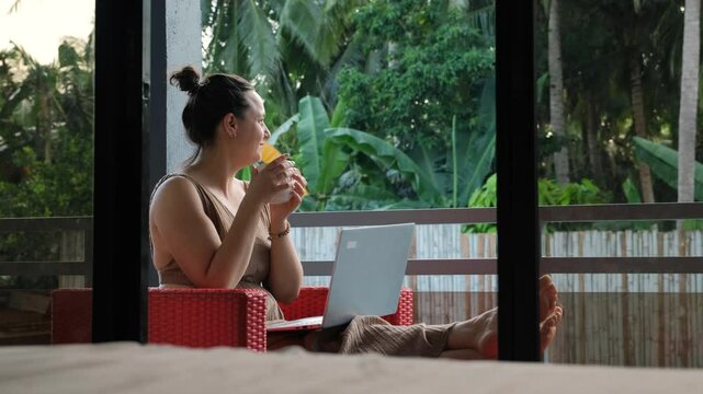 A young woman relaxes on a balcony, working on her laptop. Surrounded by lush tropical greenery, she sips a fresh morning coffee, embracing a serene atmosphere