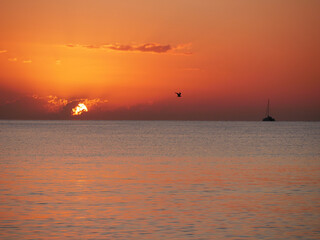 A breathtaking sunset over the calm sea with a sailboat on the horizon. Warm orange and golden tones fill the sky and reflect on the water. Travel, nature, relaxation, and maritime concept