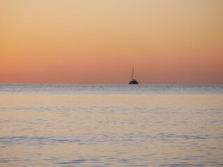 A breathtaking sunset over the calm sea with a sailboat on the horizon. Warm orange and golden...