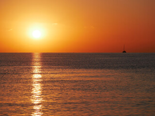 A breathtaking sunset over the calm sea with a sailboat on the horizon. Warm orange and golden tones fill the sky and reflect on the water. Travel, nature, relaxation, and maritime concept