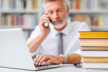 Fototapeta premium Professional man in a white shirt using a laptop while talking on the phone, surrounded by stacks of books, illustrating a busy office environment with focus on multitasking