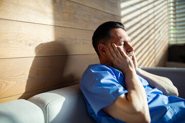 Tired male nurse sitting on couch with hands on face in sunlight