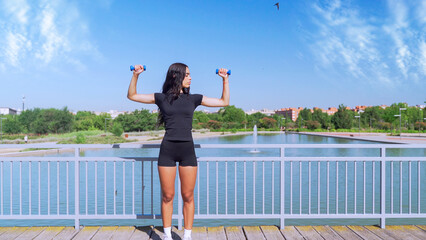 Fitness model exercising with dumbbells by a lake in a city park, promoting healthy lifestyle and urban fitness