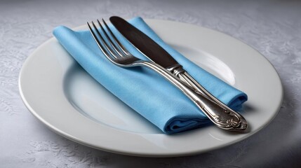 White plate with a blue napkin folded neatly on top of it. on the plate, there is a silver fork and knife with a curved handle. the plate is placed on a gray tablecloth with a floral pattern.