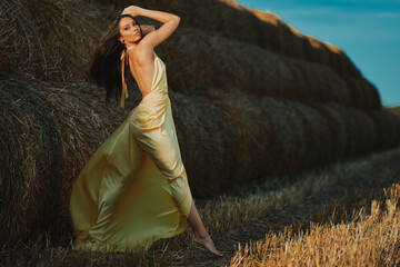 Fashion model posing in golden dress near hay bales at sunset