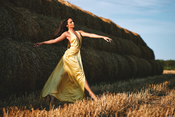 Young woman dancing barefoot in a yellow dress near hay bales in a field at sunset