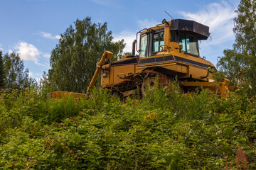 Yellow bulldozer on grassy hill surrounded by wild plants and trees under a bright sky. Concept of...
