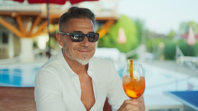 Handsome mature man smiling while holding cocktail by poolside on sunny summer vacation day. Portrait of happy, content male in sunglasses sitting in hotel resort garden on holiday trip.