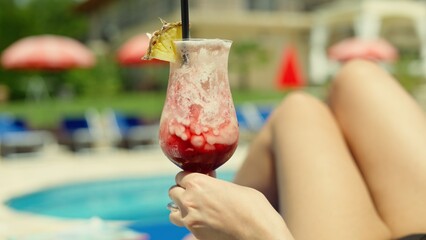 Female hand holding refreshing cocktail by pool on a sunny summer day. Woman enjoying typical dream...