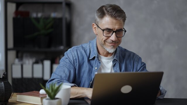 Fototapeta Mature entrepreneur with glasses concentrating on work, typing on laptop in office. Busy professional man, business owner. Middle aged businessman managing startup company, making financial decision.