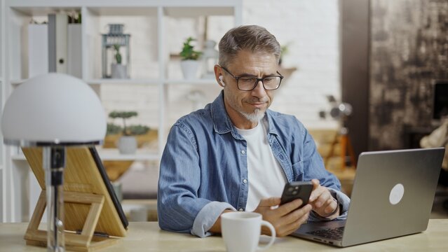 Middle aged man working in home office, using smartphone, laptop. Businessman sitting at desk in apartment to remote managing business. Freelancer entrepreneur checking messages on phone.