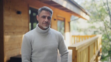 Portrait of confident senior man with grey hair outdoor on home balcony. Mature businessman on retreat standing on the porch of wooden mountain cabin, enjoying tranquility and success of hard work.