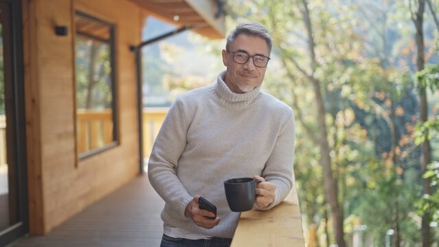 Relaxed man drinking coffee using mobile phone on balcony of modern wooden house in mountains. Portrait of happy mature businessman enjoying nature and peaceful moment of vacation, smiling. - Powered by Adobe