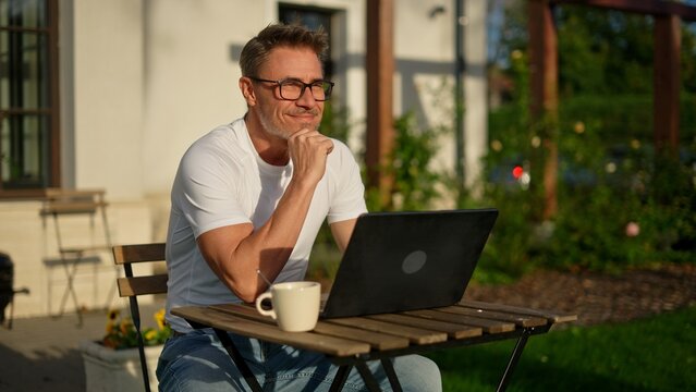 Happy businessman working remotely in garden. Mature man using laptop computer outdoor in morning with coffee. Male entrepreneur freelancer tele working online in home office. Digital business life.