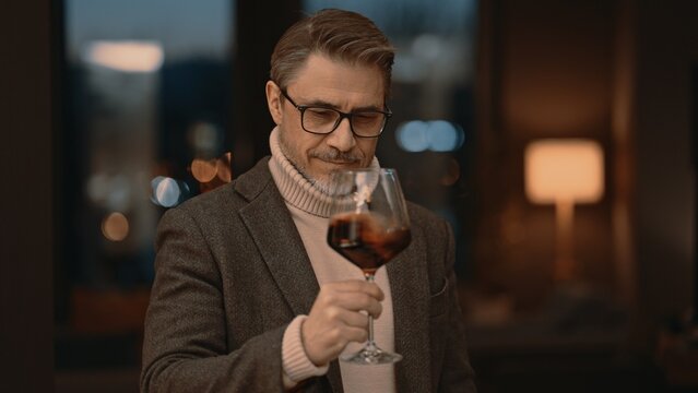 Elegant mid adult man drinking, tasting red wine from glass at home in cosy room. Portrait of happy, satisfied middle aged man, smiling.