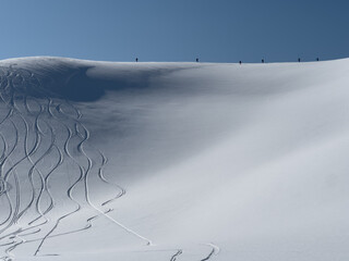 Skiers ascend a snowy ridge under a clear sky, with fresh powder tracks carving elegant lines below. A perfect day in the backcountry.