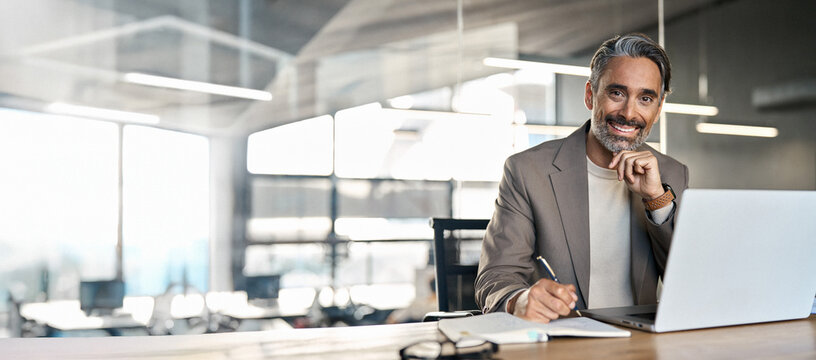 Happy middle aged professional business man, older executive ceo manager, smiling mature entrepreneur lawyer wearing suit sitting at office desk working on laptop computer. Copy space. Portrait.