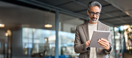 Busy mature older business man executive standing in office using digital tablet. Middle aged...