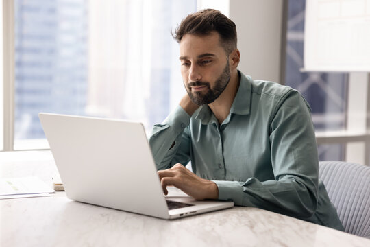 Focused man sitting at desk review information received in e-mail, learns new business AI platform, web surfing using laptop in modern high-rise building office. Innovative tech, business, workflow