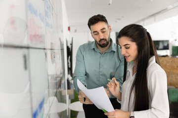 Teammates collaborating on paperwork standing next to white board, reviewing project timeline,...