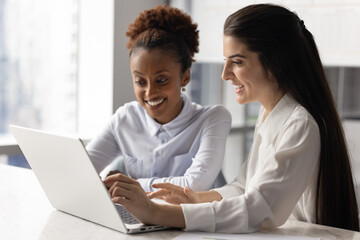 Young African and Latina women teammates engaged in teamwork seated at desk in modern workplace...