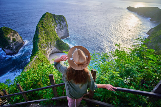 Woman traveler stands on steps of stairs and enjoying landscape of scenic Kelingking beach during vacation to Nusa Penida Island in Indonesia, travel to touristic beautiful destination