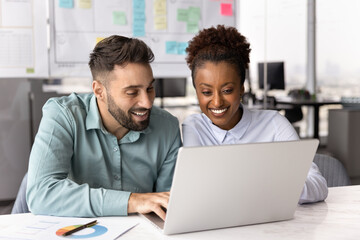 Two multinational smiling colleagues looking at laptop screen seated together at desk in modern...