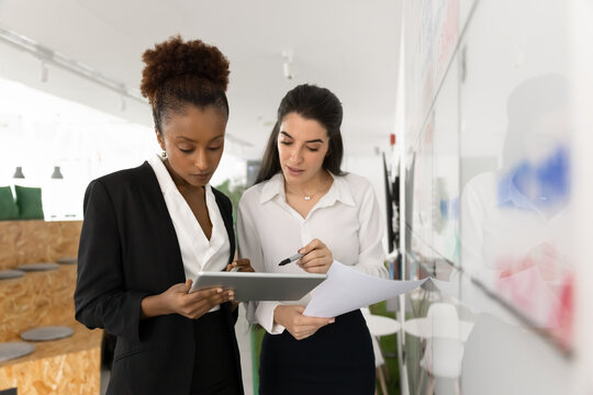 Two attractive focused women workmates reviewing information displayed on tablet, discussing data or content shown on screen, collaborating on project standing next to white board in modern coworking