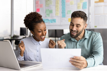 Two happy workmates sit at desk read document, getting approval for proposal or idea, receiving positive feedback or recognition for their work, closing deal, celebrating team achievement and success