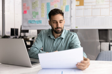 Young Hispanic professional man sitting at table with open laptop, reviewing financial report or...