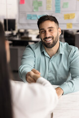 Young Hispanic businessman shaking hands with woman client seated at desk in meeting room, vertical...