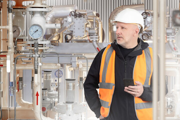 Man in boiler room. Gas equipment behind worker. Boiler unit with pipes. Man engineer in orange...