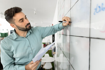Young focused man engaged with large, gridded whiteboard, holding papers and marker, writes...