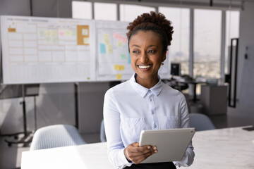 Smiling African woman, office employee standing in modern meeting room with tablet, look into...