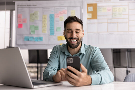 Cheerful man use smartphone, enjoy personal chat exchanges messages through mobile application, learn new AI platform for business or fun, distracted from workflow on laptop in workplace. Modern tech - Powered by Adobe