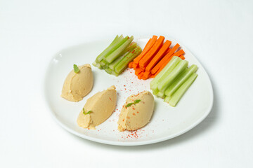Plate of hummus with fresh vegetables — carrot, celery, and cucumber sticks — on a white background